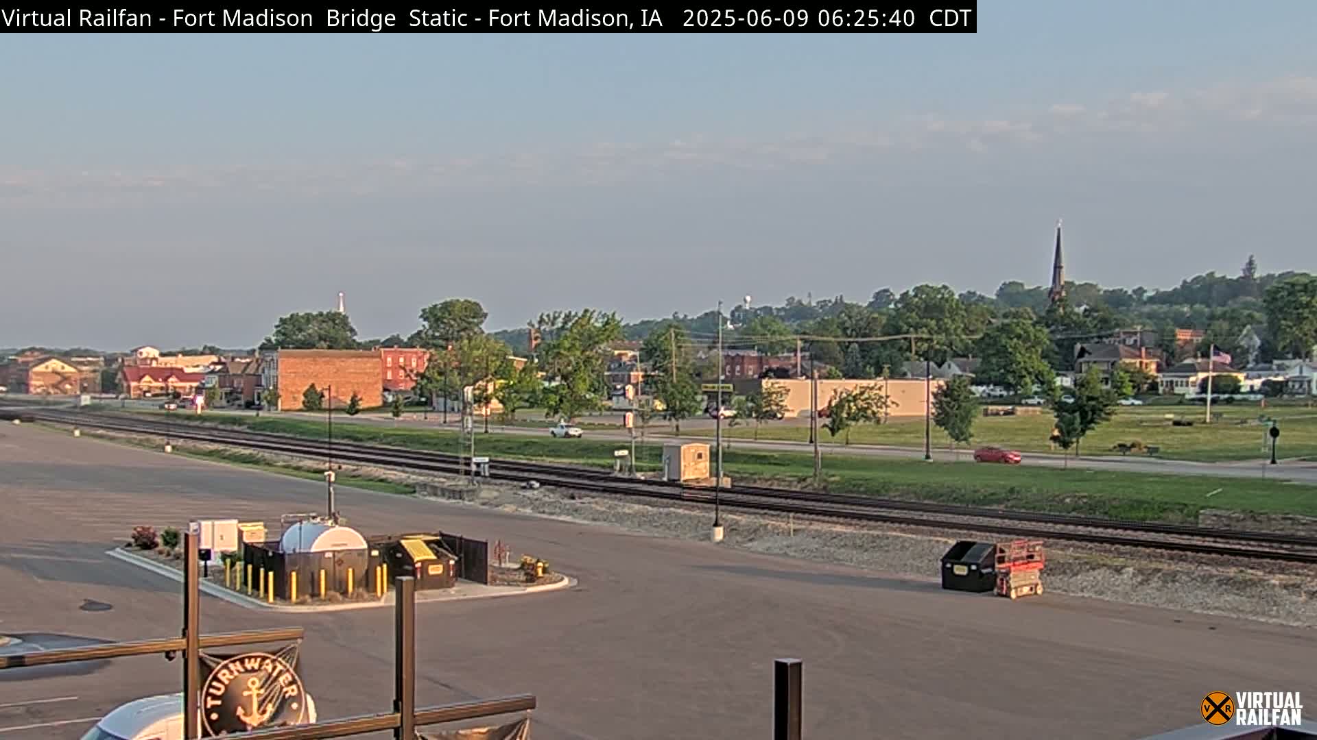 A long shot of a town under a mostly clear sky, showing railroad tracks, a parking lot, and several buildings, with a few cars visible on the road.