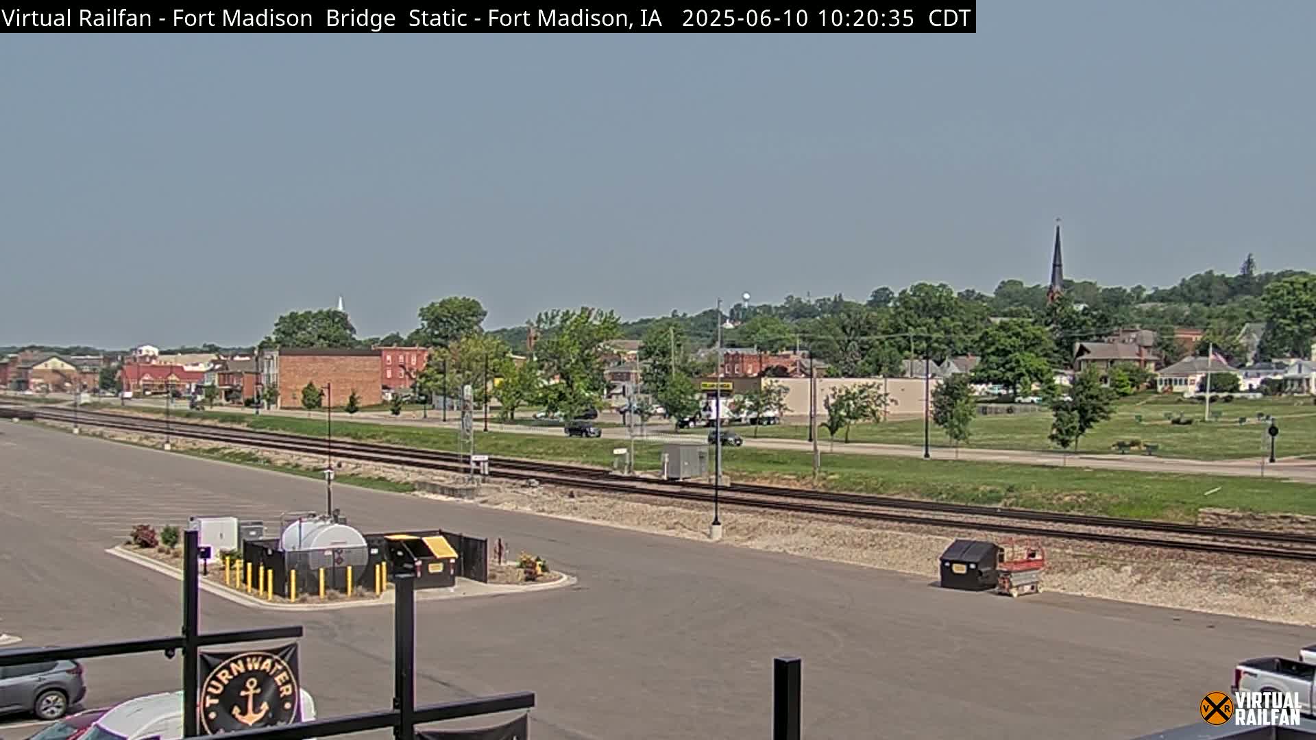 A small town with brick buildings and a tall church steeple is visible beyond railroad tracks on a mostly sunny day.
