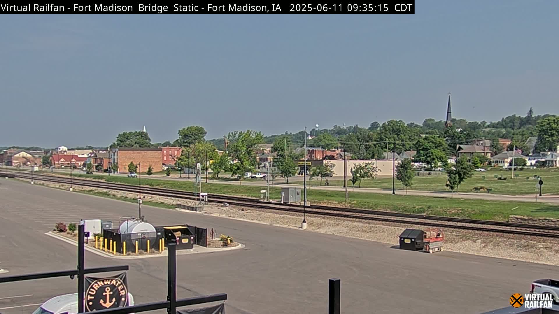 A view of a town under a clear sky, showing railroad tracks in the foreground and buildings in the midground, with a large grassy area separating the tracks from the buildings.
