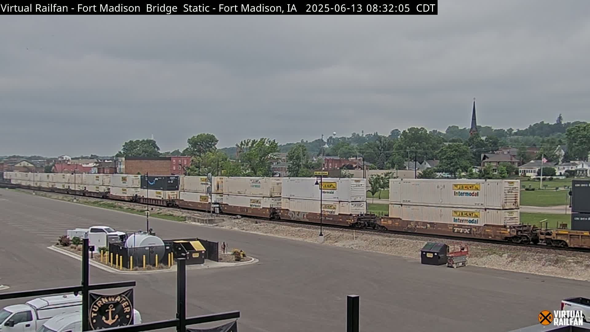 A long freight train carrying numerous stacked shipping containers is visible, with a small town and overcast sky in the background.