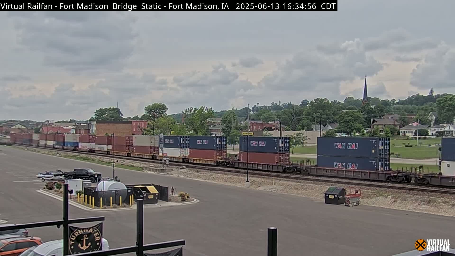 A long freight train carrying multiple shipping containers travels along railroad tracks under a partly cloudy sky, with a small town visible in the background.