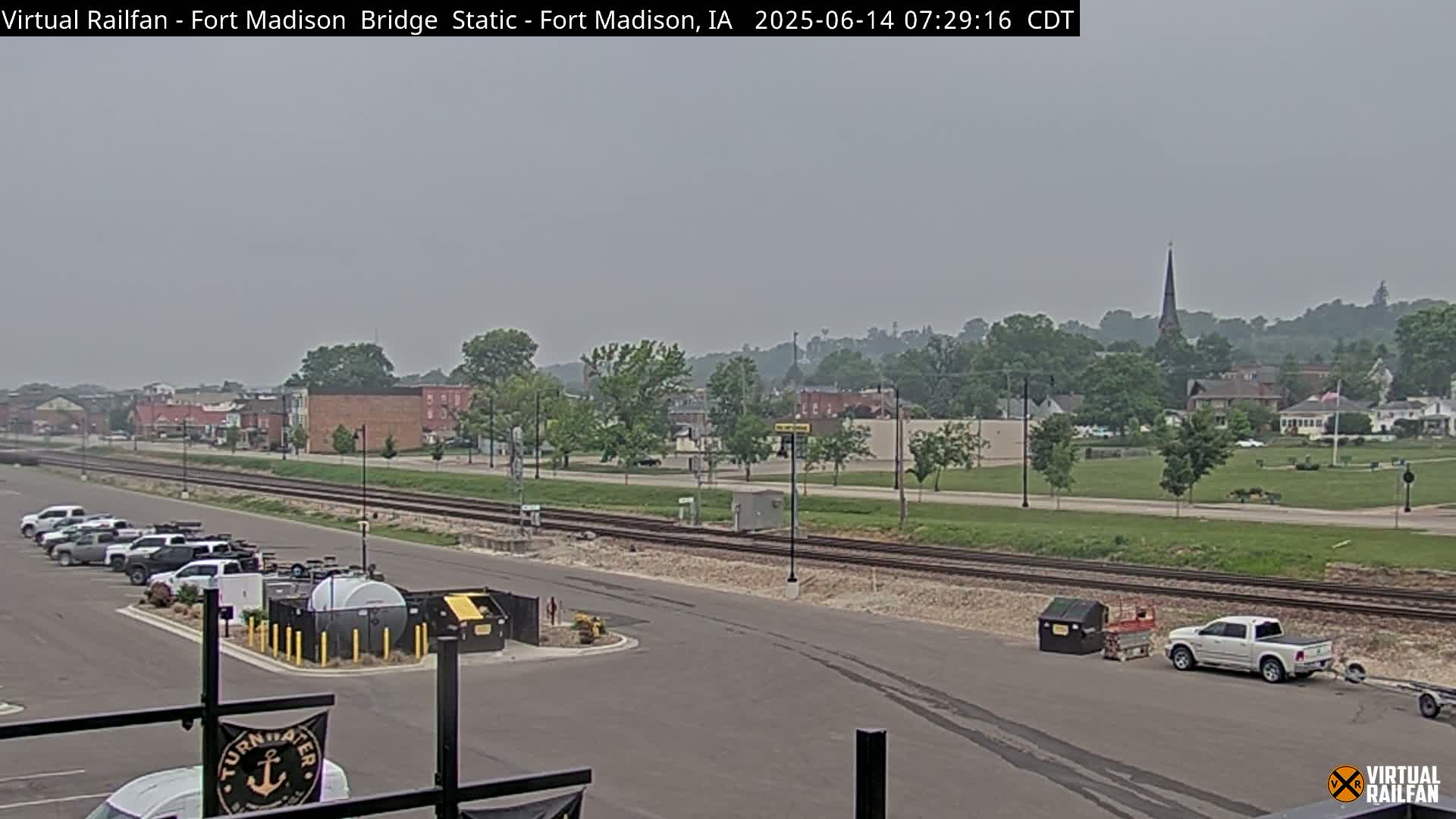 A hazy view of a town, railroad tracks, and parked vehicles, with a church steeple visible in the distance.