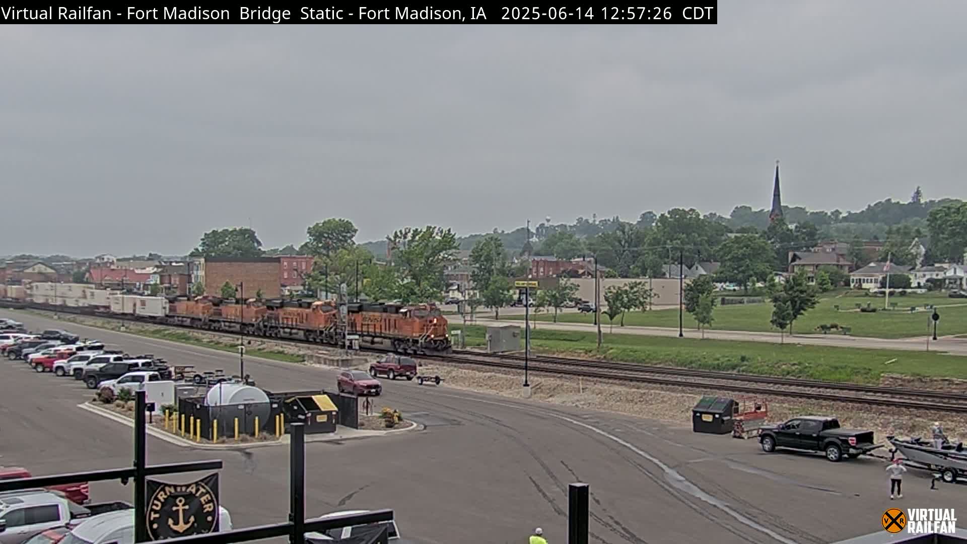 A long freight train passes by a parking lot and town under an overcast sky.