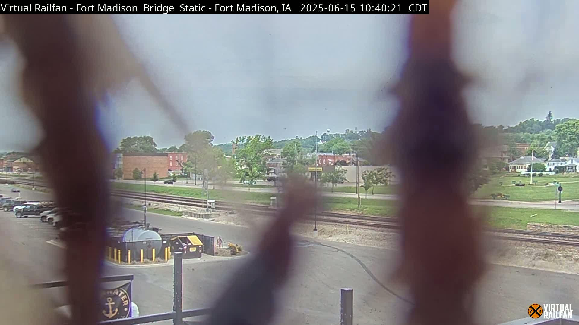 A view of a town, seen through a fence, on an overcast day, shows railroad tracks, buildings, vehicles, and a parking lot.