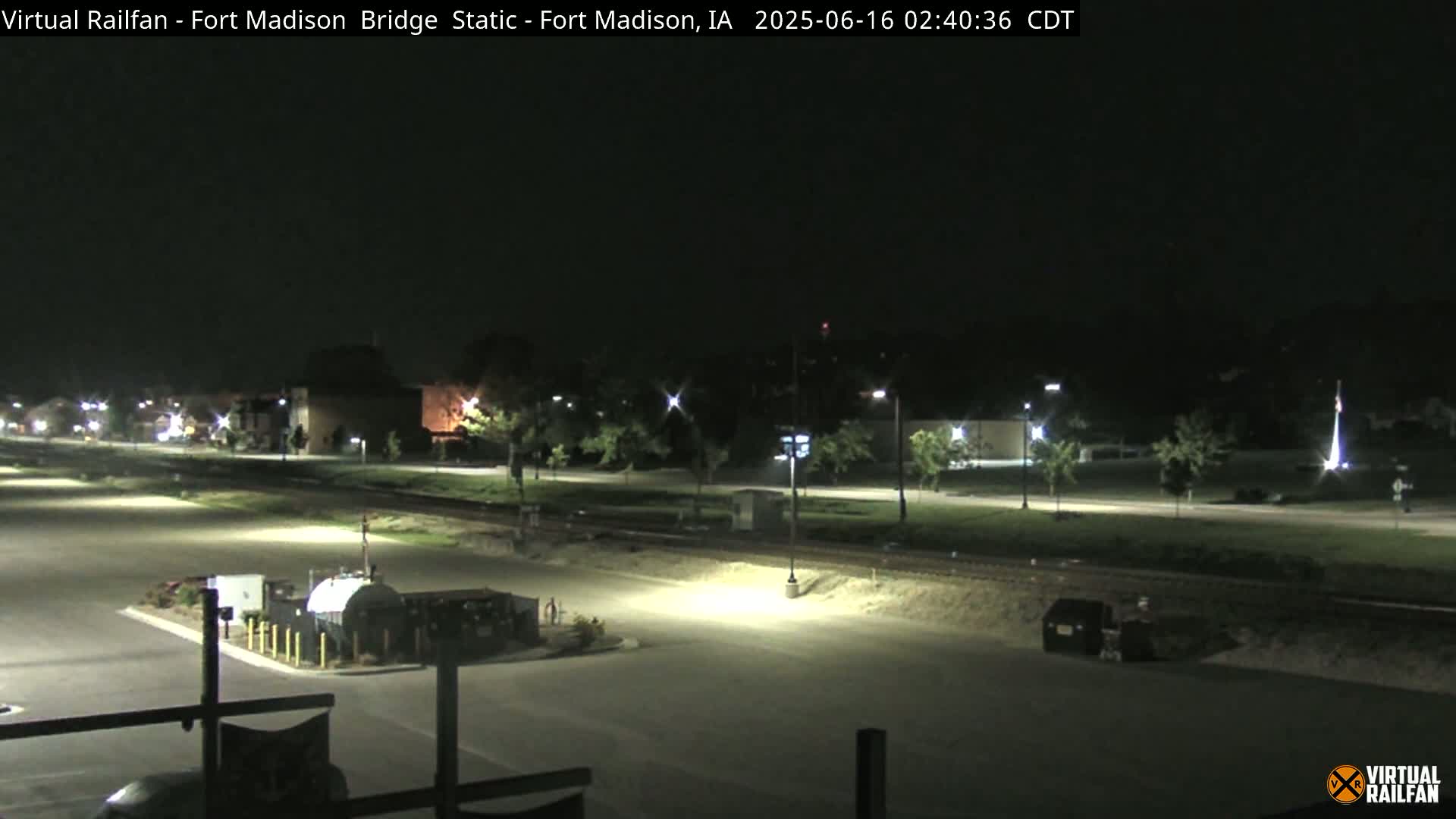 A nighttime view of a mostly empty parking lot and roadway next to a railroad track, with some buildings and trees in the background under a clear, dark sky.