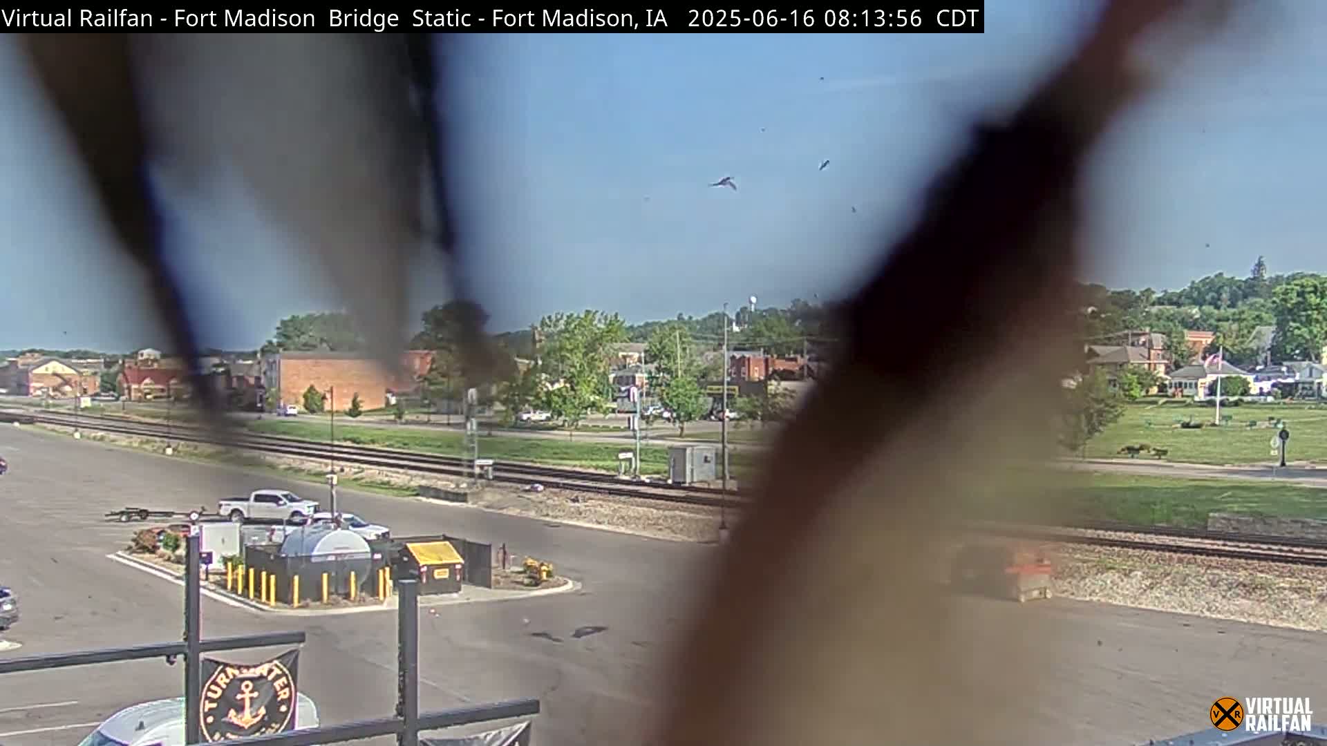 A partly obstructed view shows a parking lot, railroad tracks, and a small town under a mostly clear sky with several birds in flight.