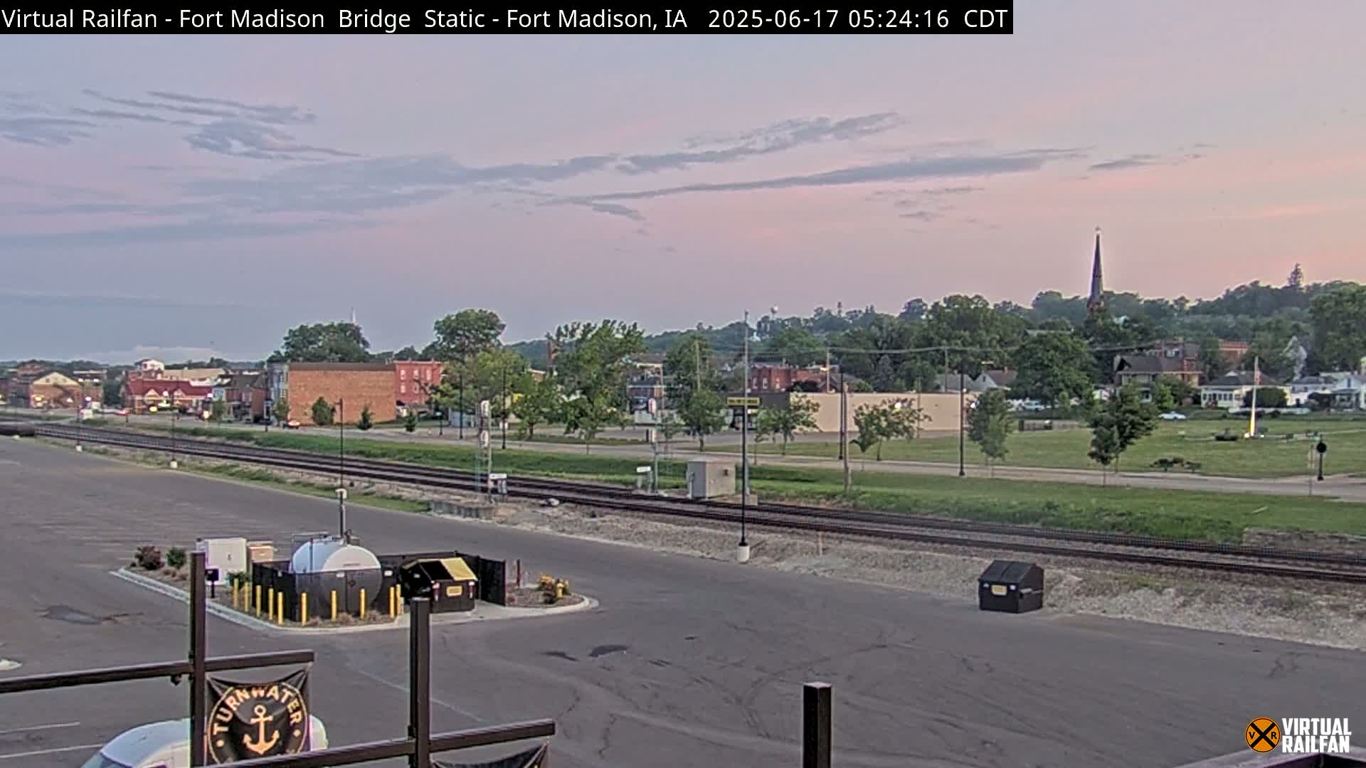 A twilight view of a town with railroad tracks in the foreground, under a pale pink and gray sky.