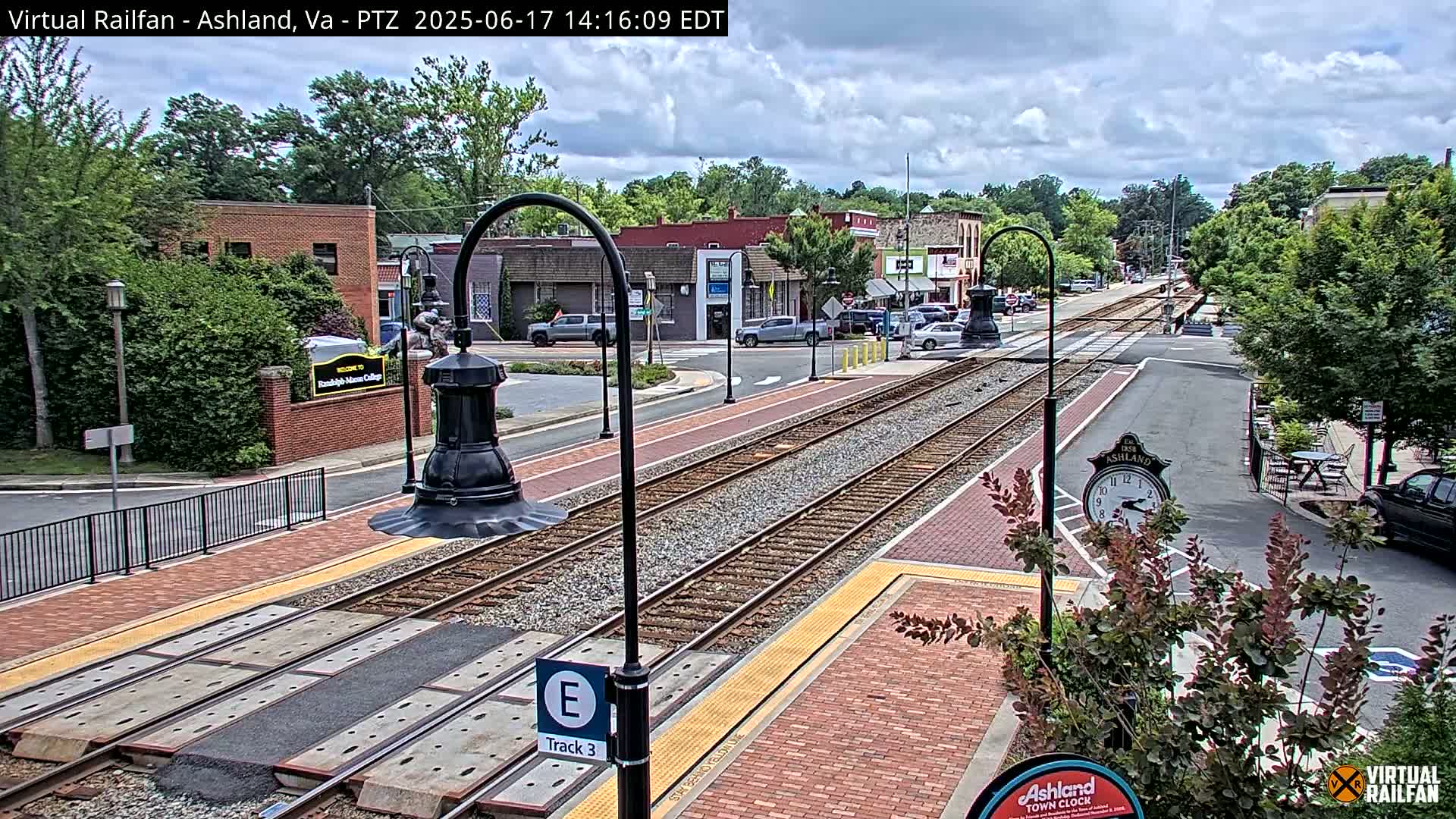 A partly cloudy day shows a train track running through a town's brick-paved streets, lined with buildings and trees.