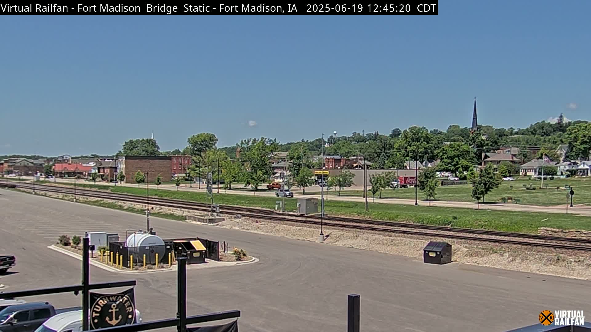 A sunny day reveals a small town with a railroad track running through it, alongside a parking lot and various buildings.