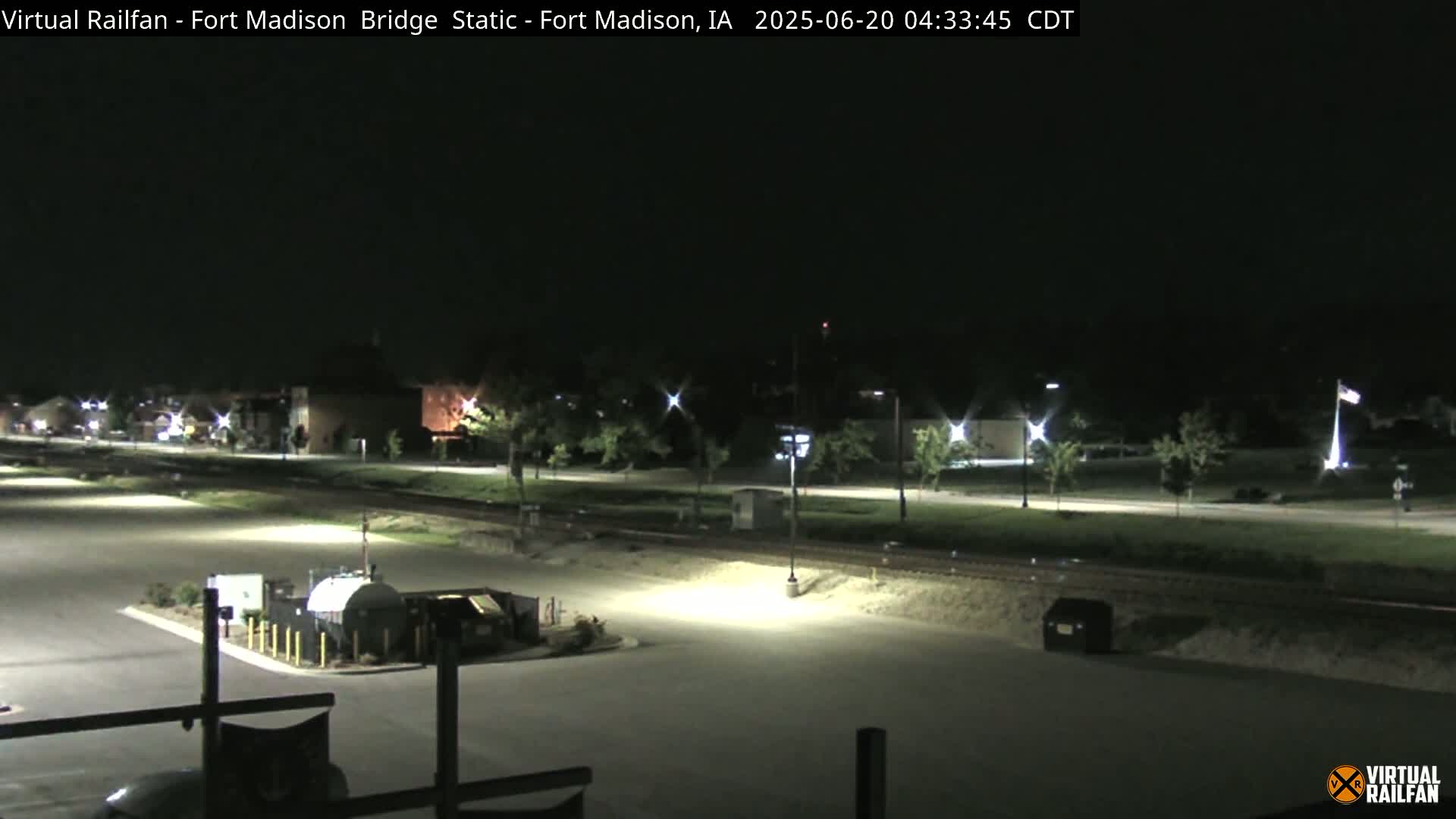 A nighttime view of an empty parking lot and roadway near a railroad track, illuminated by artificial lights.
