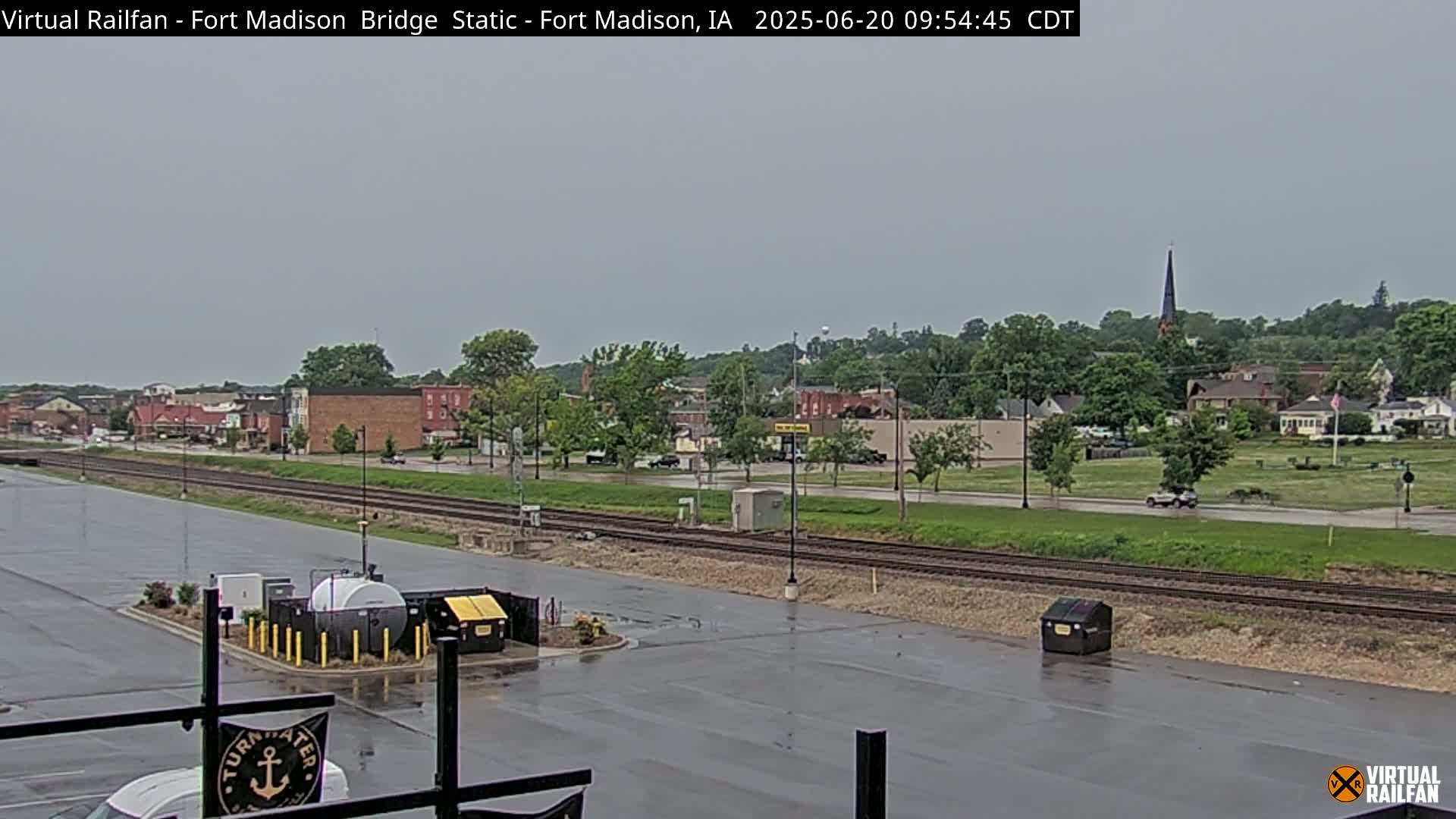 A wet, overcast view of a town's railroad tracks and buildings, seen from a parking lot.
