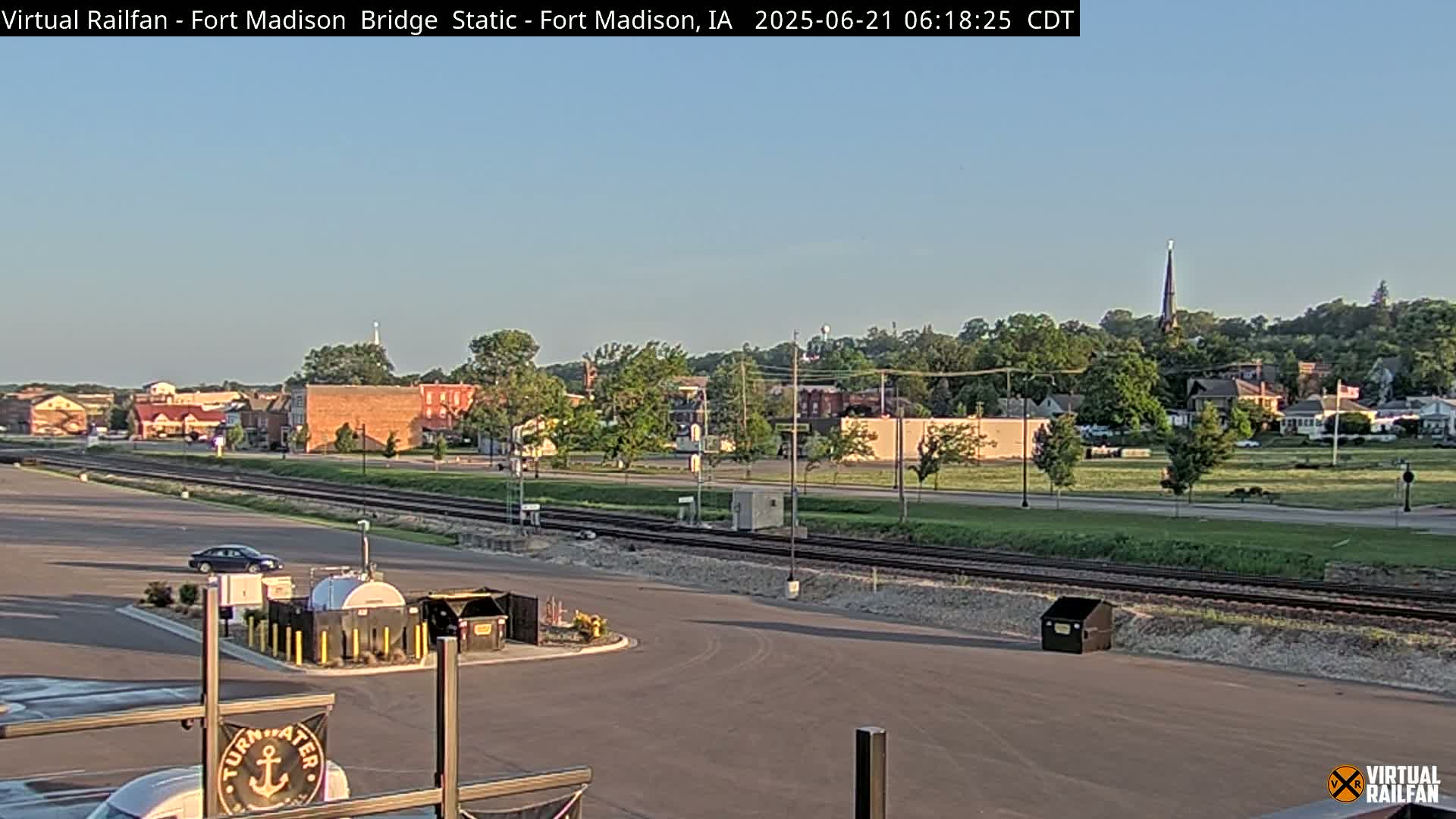 A clear, sunny morning view of a town's outskirts, featuring railroad tracks and a parking lot with some industrial-looking containers.