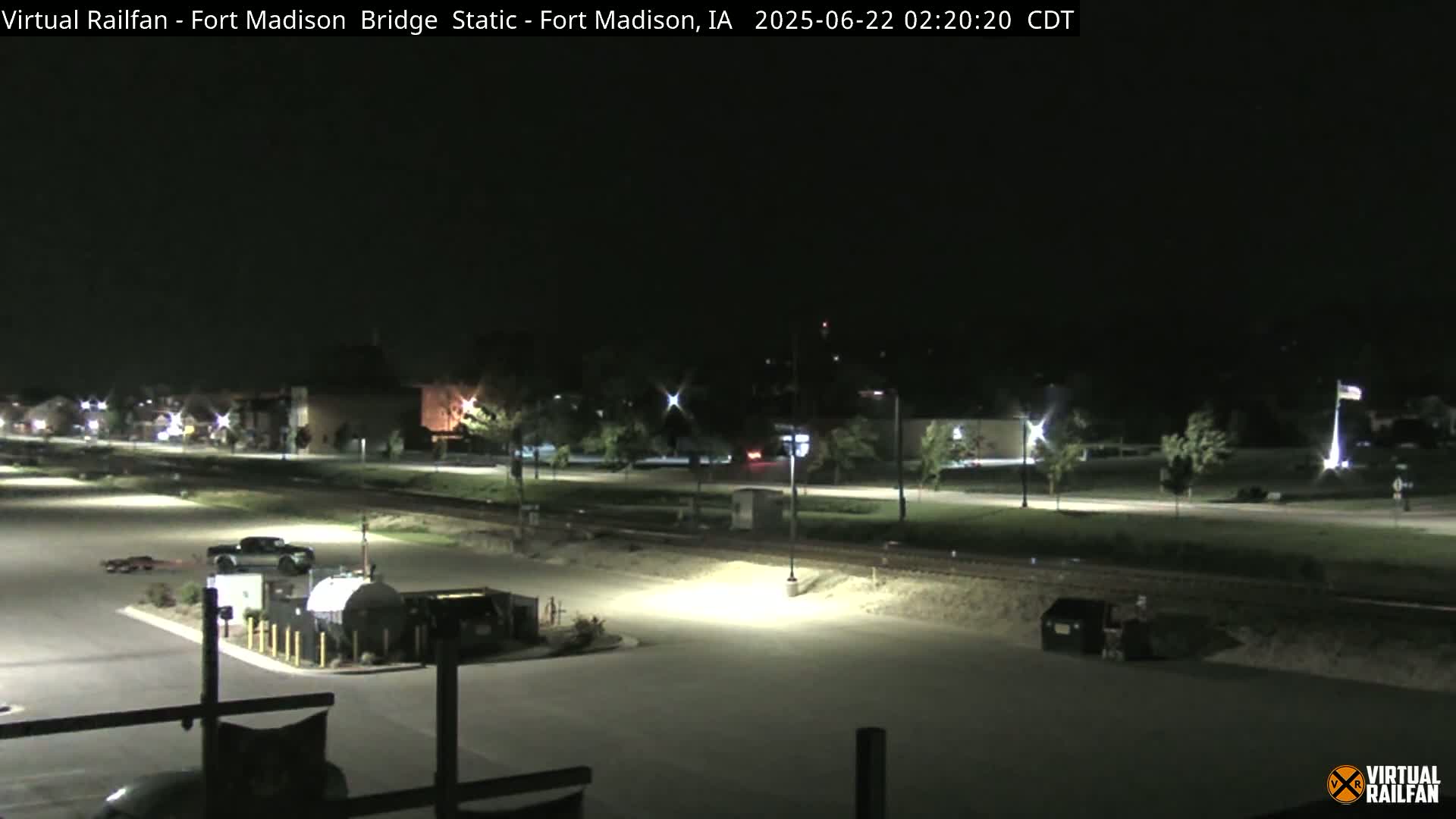A nighttime view of a mostly empty parking lot next to a road and railroad tracks, with a few vehicles visible and some buildings in the background under a clear, dark sky.