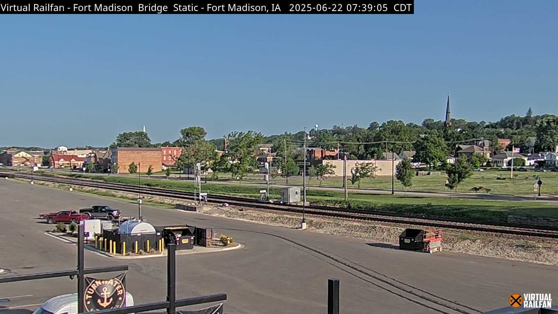 A clear, sunny day reveals a small town with a railroad track running through a parking lot, with a few vehicles and storage containers visible.