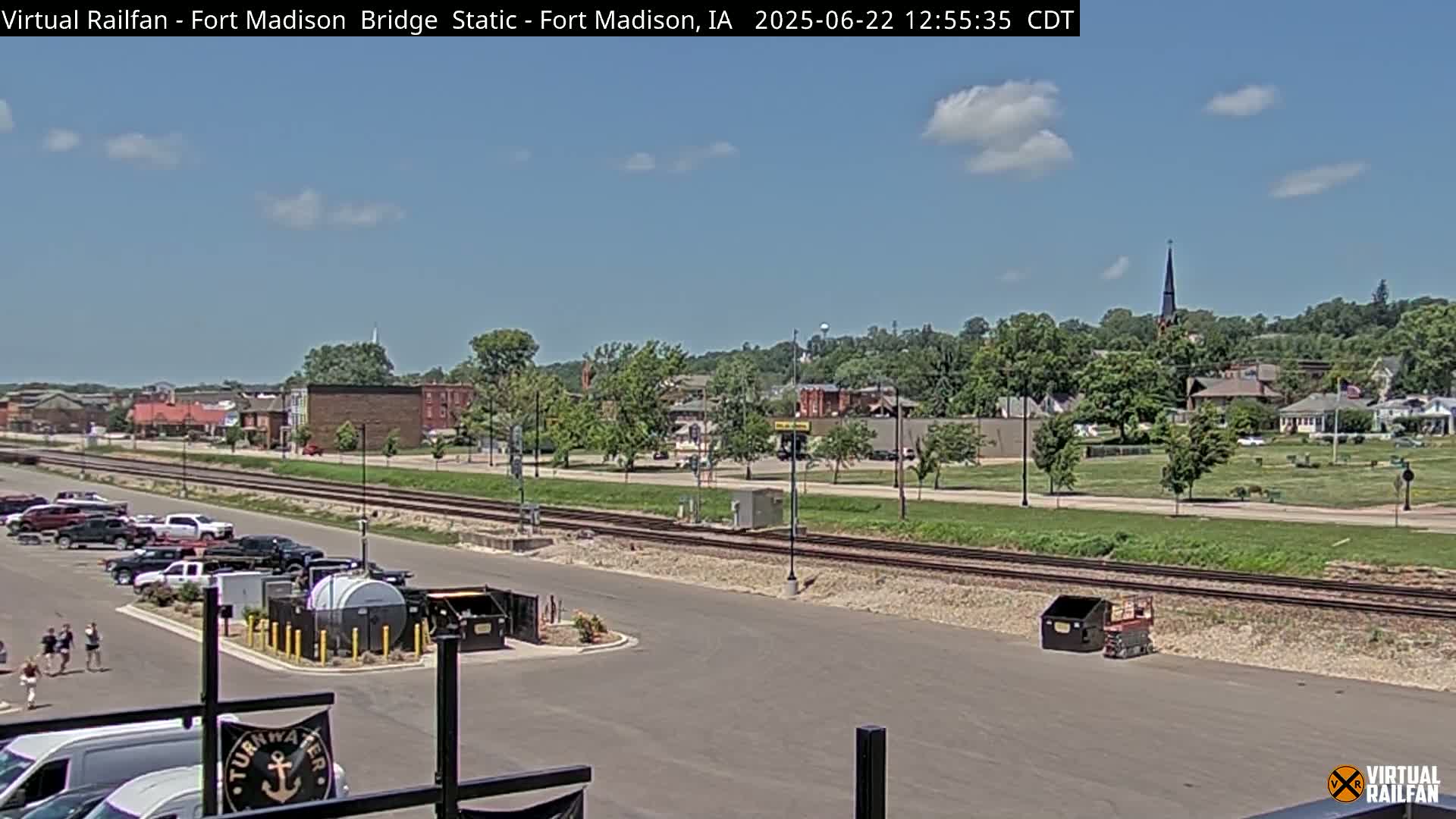 A mostly sunny day reveals a view of a town with train tracks running alongside a parking lot, with a few people and vehicles present.