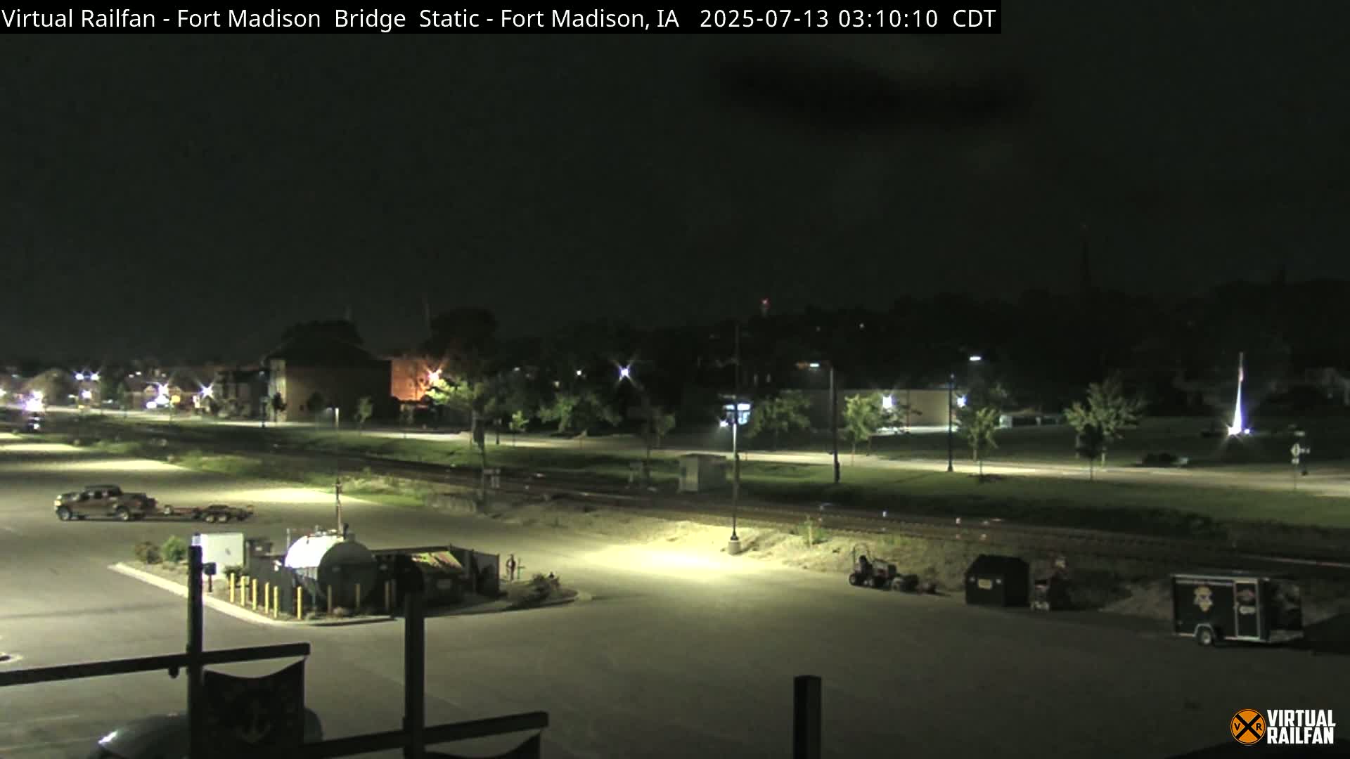 A nighttime view of a mostly empty parking lot, a roadway, and some buildings under artificial light.