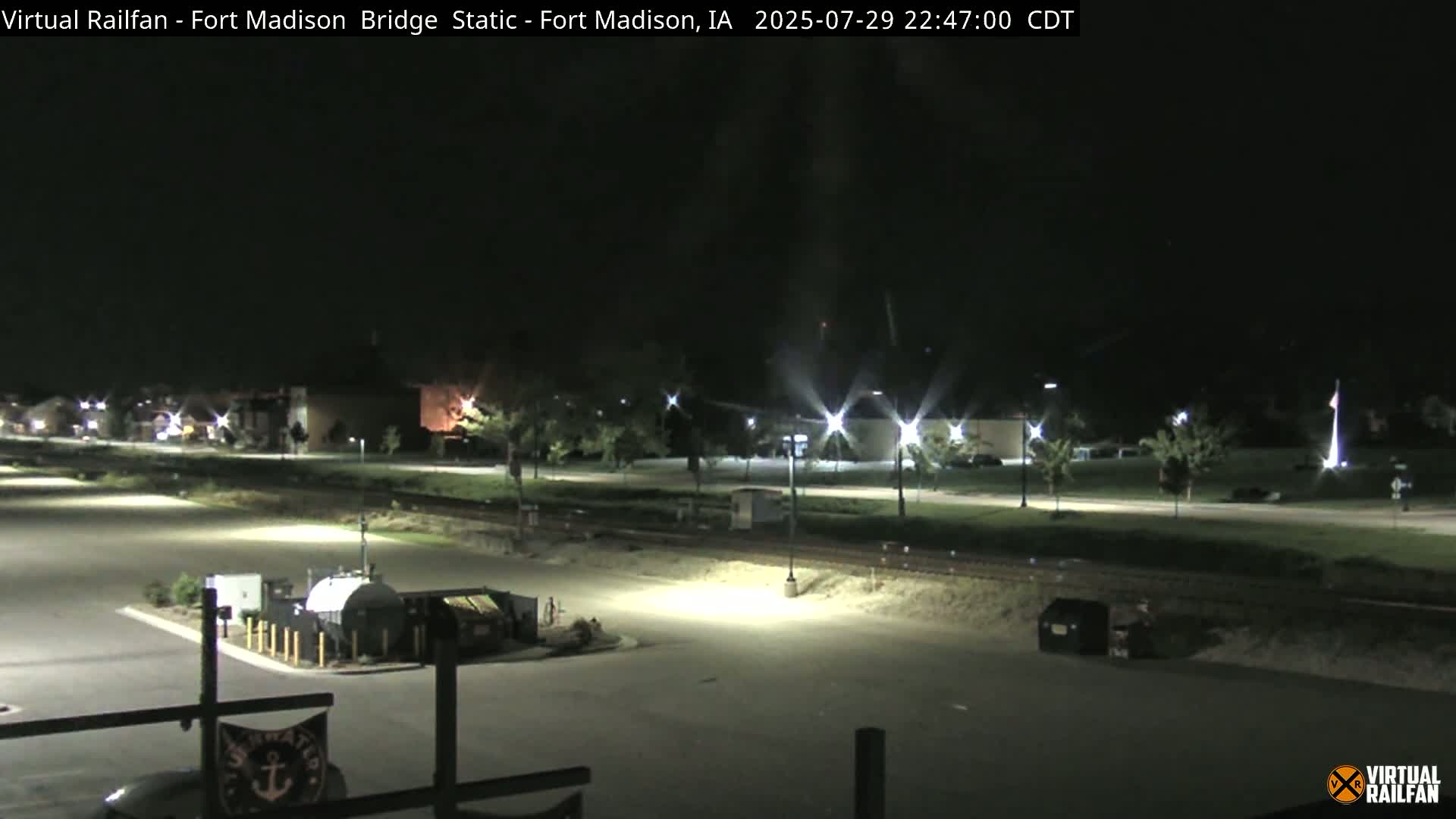A nighttime view of a mostly empty parking lot, a roadway, and some buildings under artificial light.