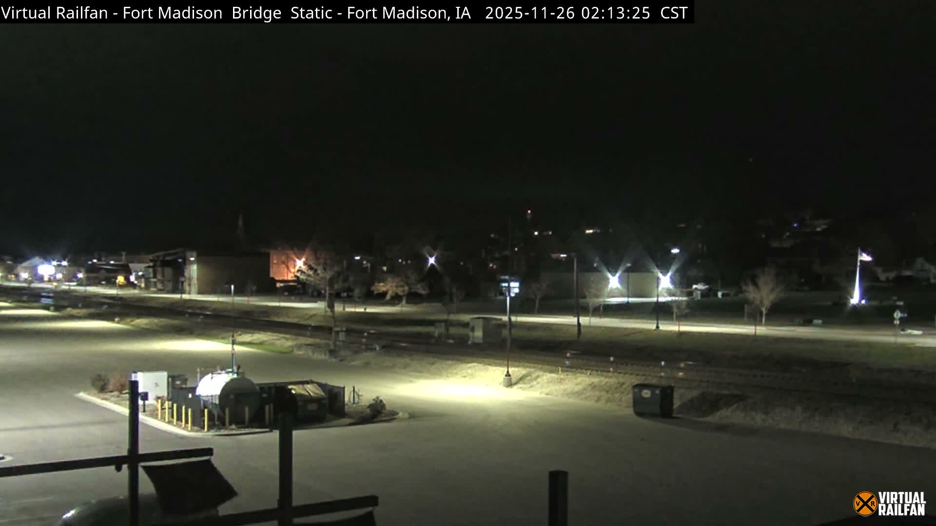 A still nighttime view reveals an illuminated commercial or industrial area featuring a large paved lot, railroad tracks, and distant buildings under a clear, dark sky.