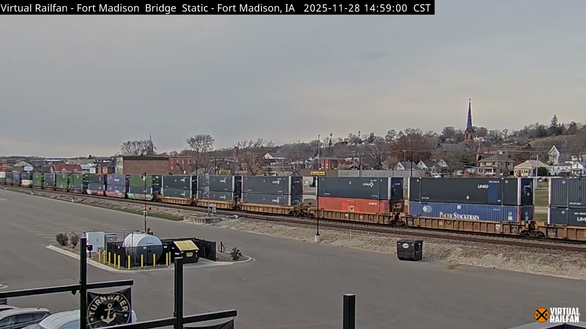 Under an overcast sky, an intermodal train laden with shipping containers stretches across tracks in the midground, with a paved area and industrial structures in the foreground, and a town featuring a prominent church spire on a distant hill.