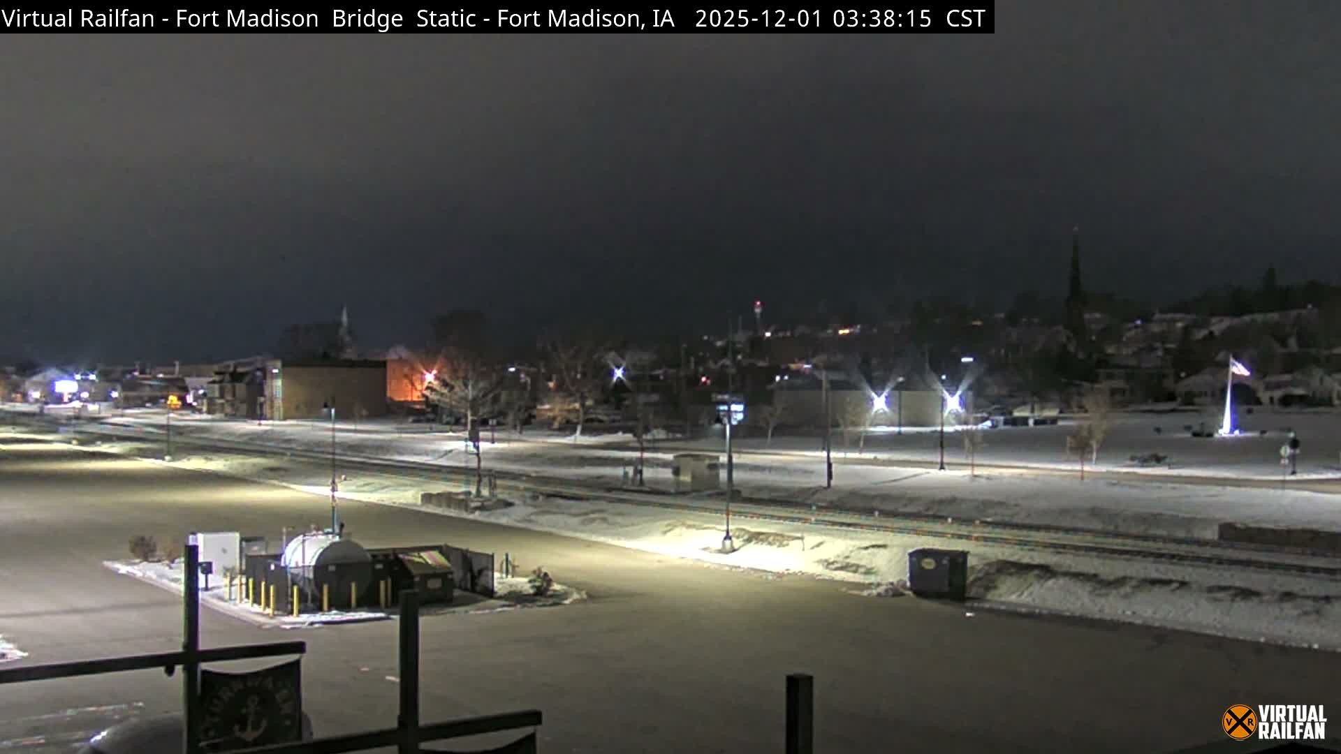 A nighttime view of a snow-covered town displays a wide, illuminated parking lot in the foreground, leading to snow-dusted railroad tracks and buildings, all under a clear, dark sky.