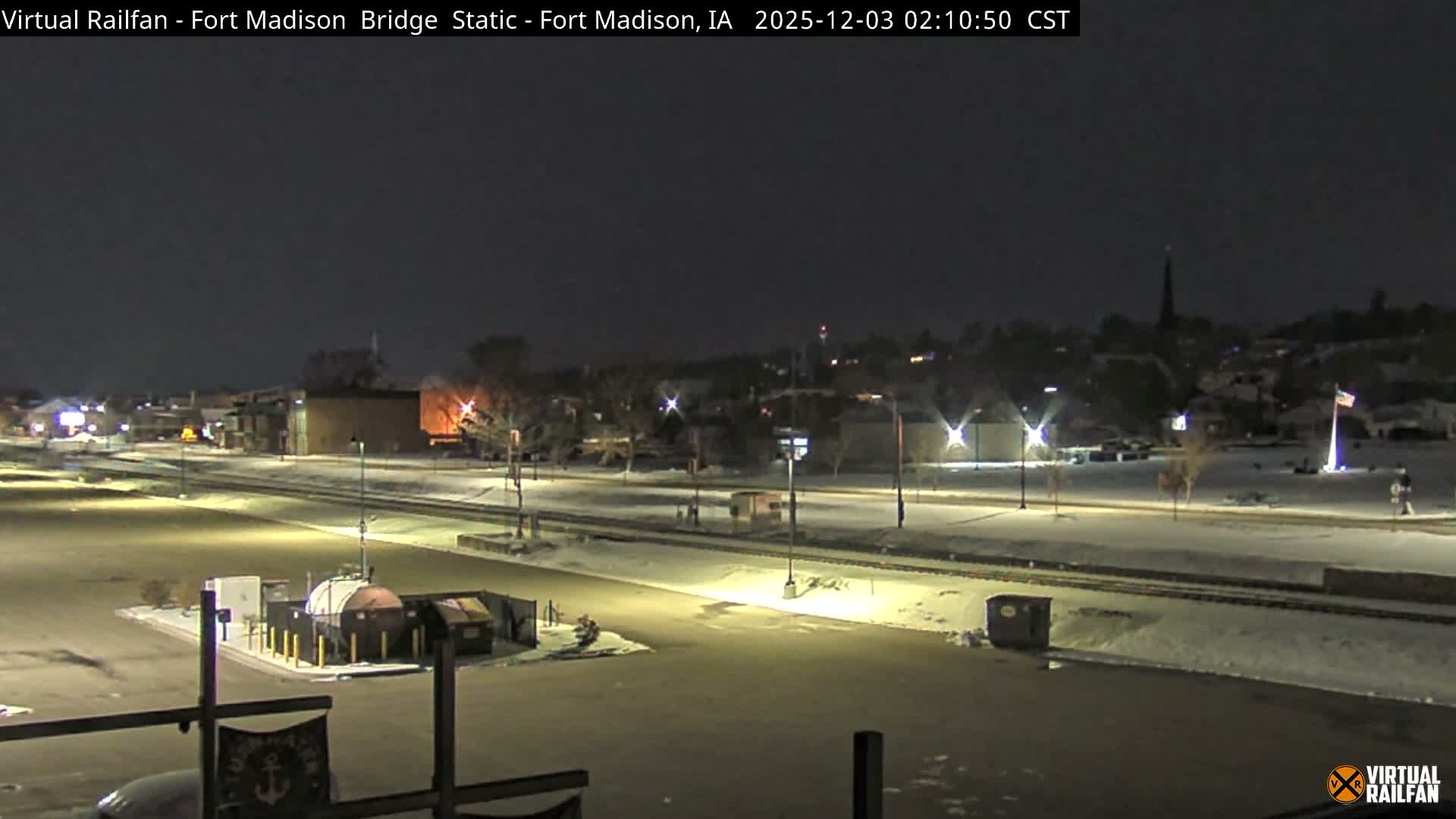 The image shows a snow-covered town at night, illuminated by streetlights, with railway tracks running through the middle ground and various buildings, including a church steeple, visible in the background under a dark sky.