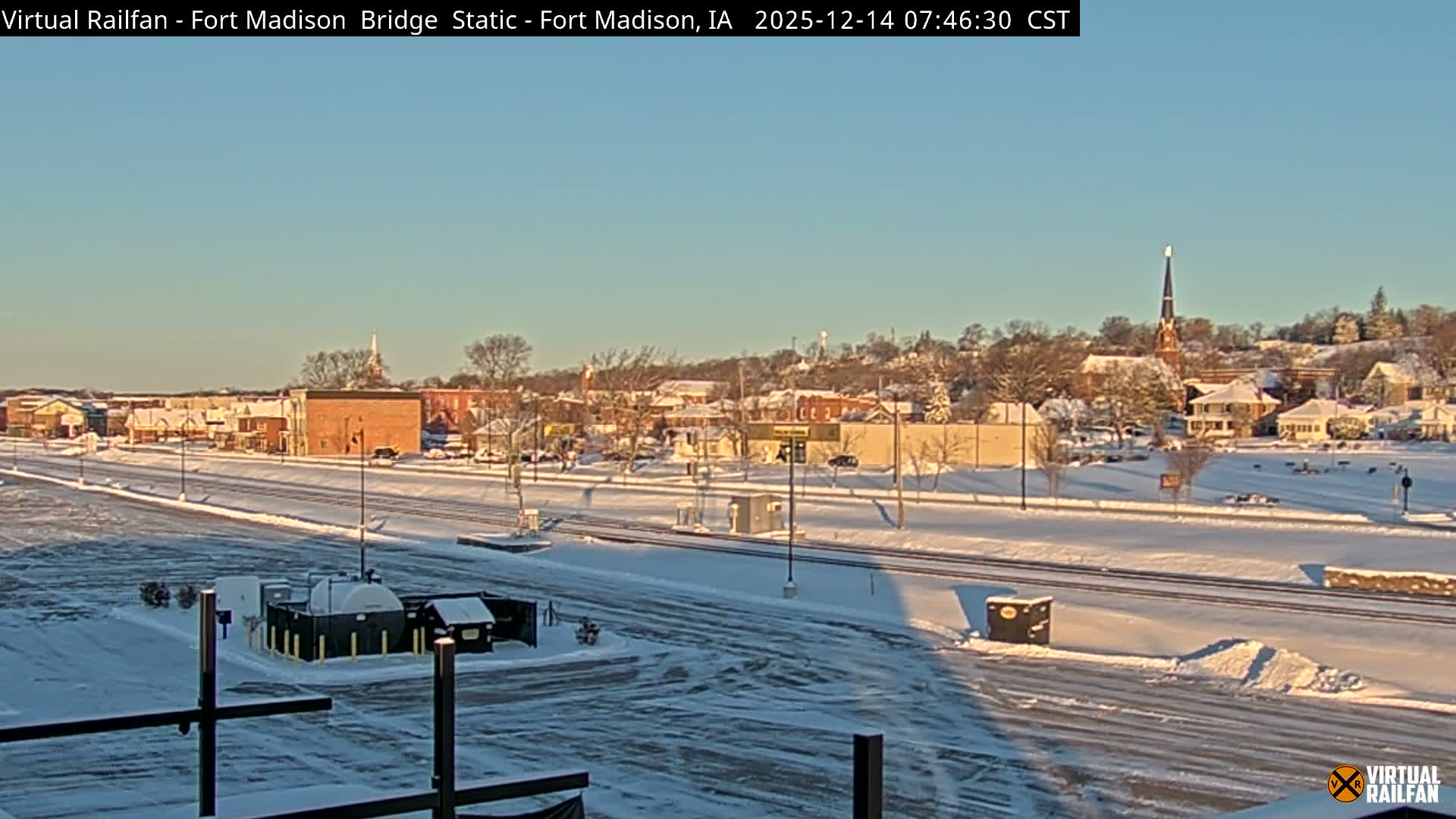 The image captures a snowy town at night, revealing a parking lot and railroad tracks in the foreground, illuminated streets, buildings, and a prominent church spire in the mid-distance, all under a dark and cloudy sky.