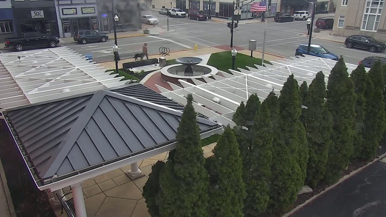 An overhead view shows a town square with a fountain, a white pergola, and a dark-roofed gazebo surrounded by evergreen trees on an overcast day.
