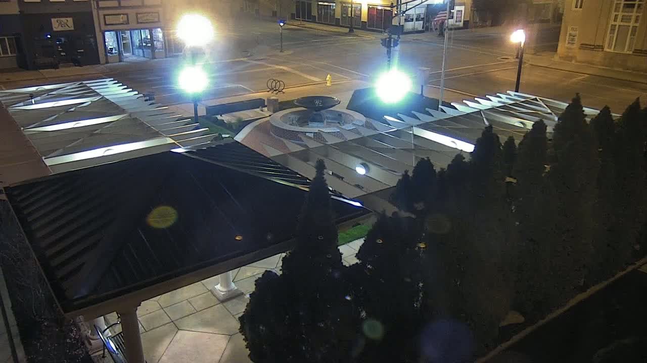 At night, a fountain is centered in a plaza surrounded by dark evergreen trees and a dark-roofed structure, brightly lit by streetlights.