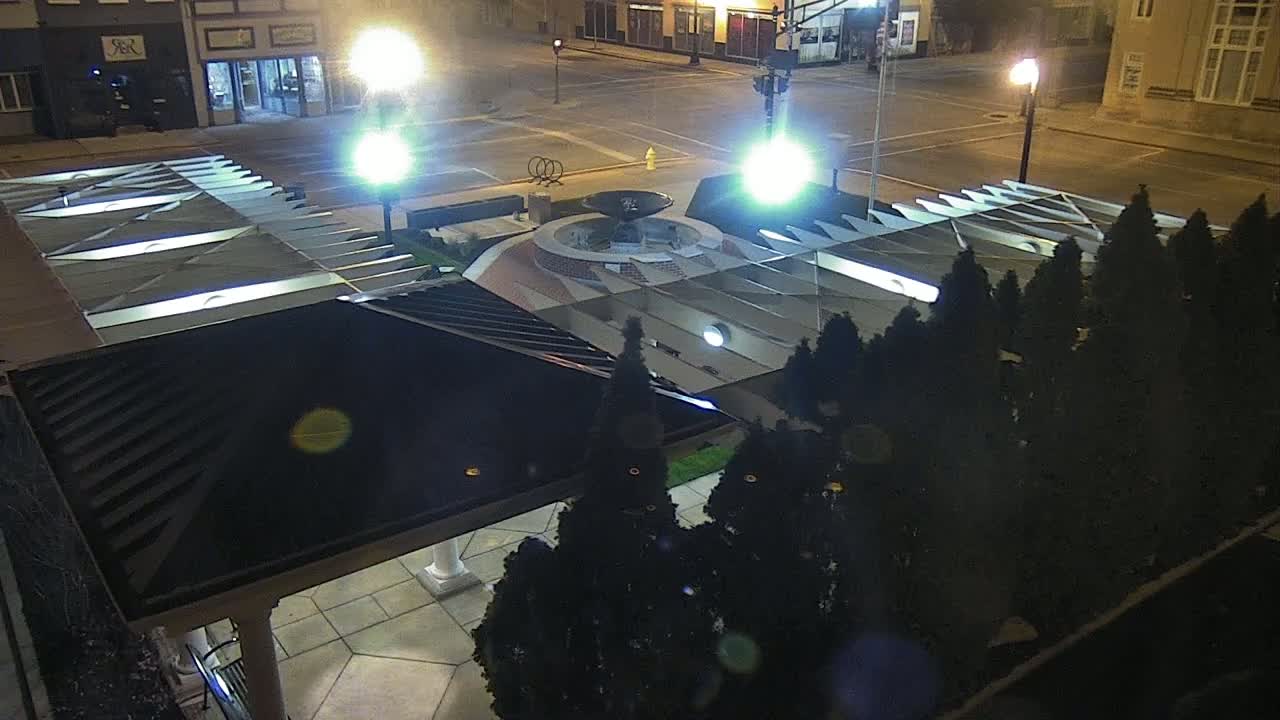 A nighttime aerial view of a town square featuring a fountain surrounded by dark evergreen trees, illuminated by streetlights.