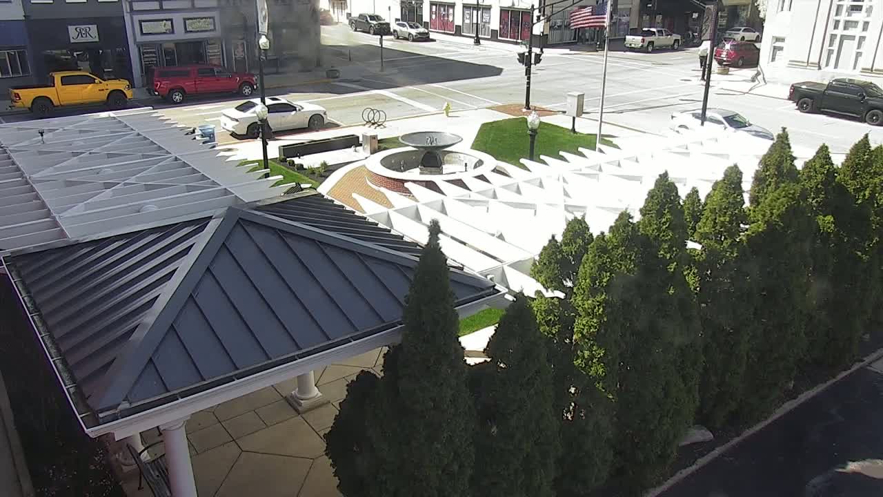 An aerial view shows a town square with a fountain, a white geometric structure, and several evergreen trees, all under a sunny sky.