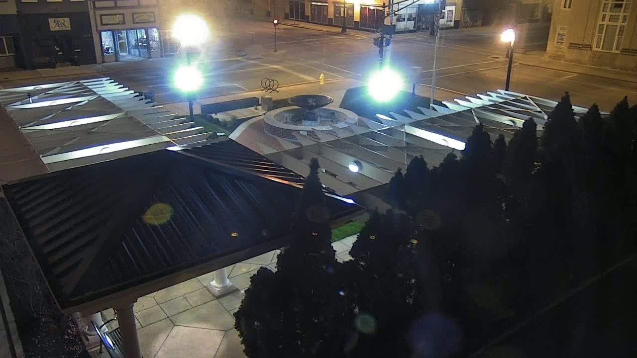 A nighttime aerial view of a plaza with a fountain at its center, surrounded by dark evergreen trees and illuminated by streetlights.