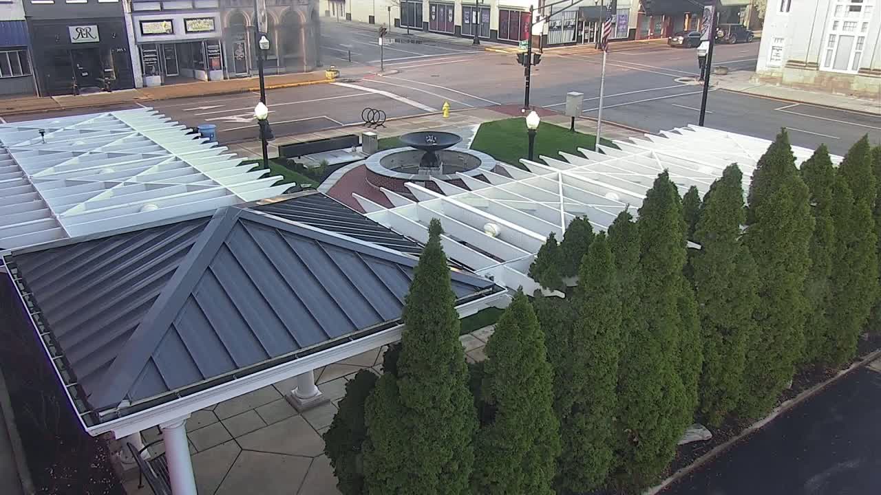 A dark gray pavilion roof sits atop a white pergola structure beside a row of evergreen trees surrounding a fountain in a town square on a sunny day.