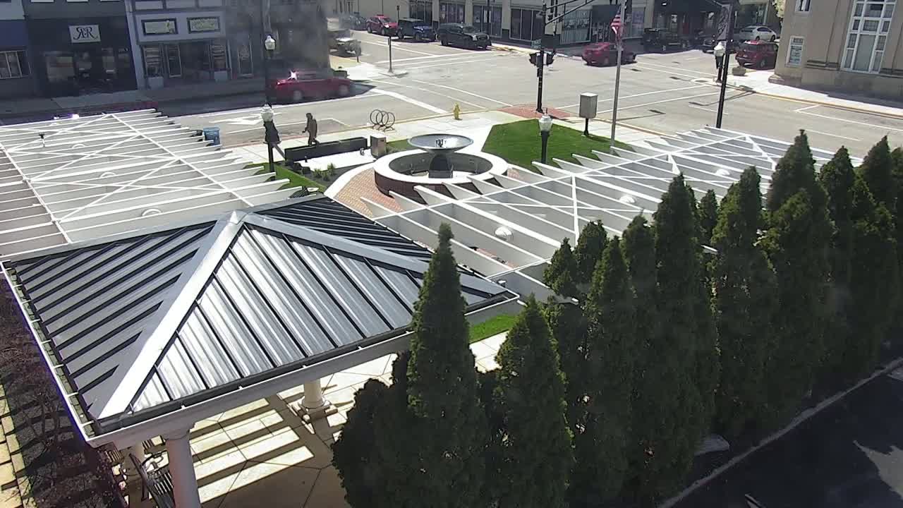 An aerial view shows a plaza with a central fountain, surrounded by evergreen trees and covered pavilions under a sunny sky.