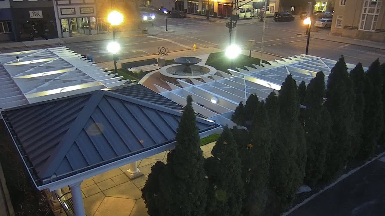 An overhead nighttime view shows a paved area with a modern pavilion, a central fountain, and a row of evergreen trees under illuminated streetlights.