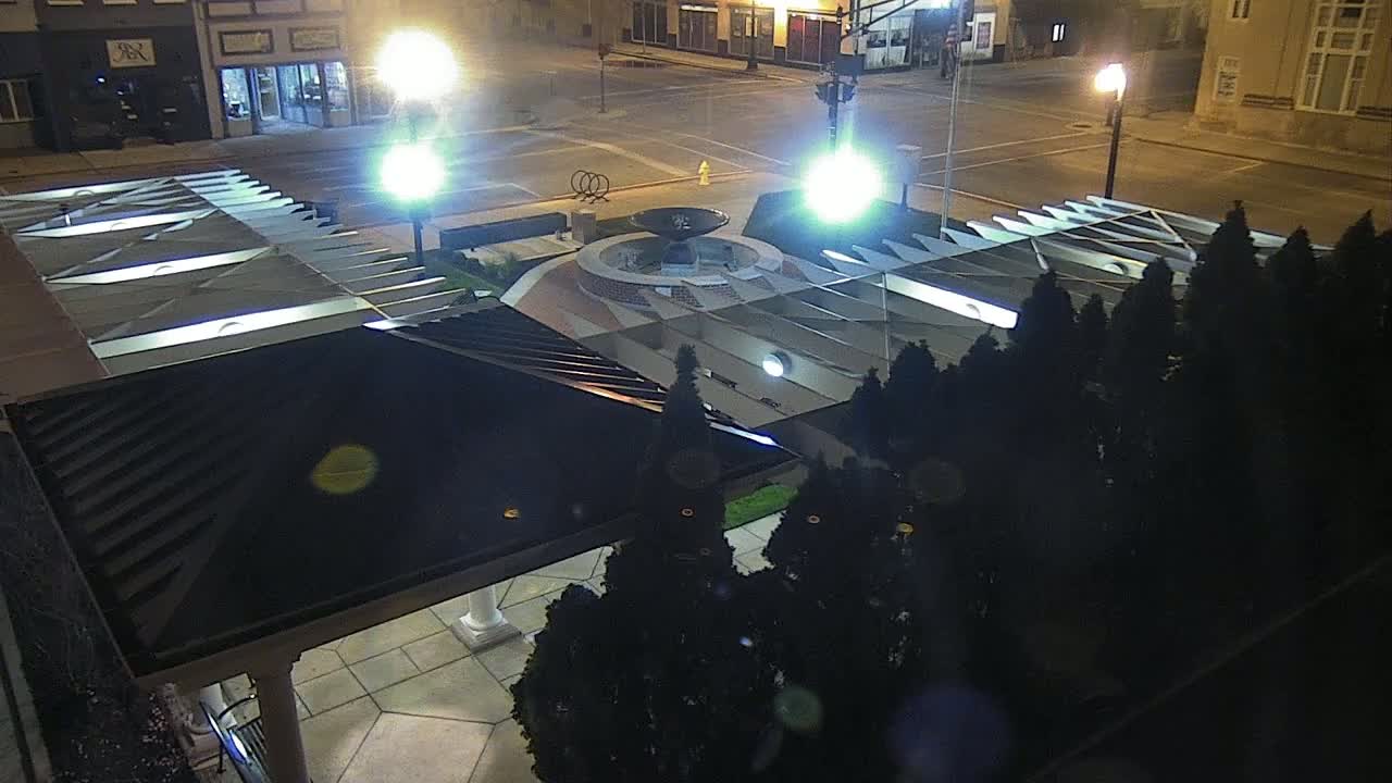 A nighttime aerial view shows a plaza with a fountain at its center, surrounded by dark evergreen trees and a covered structure, under bright streetlights.