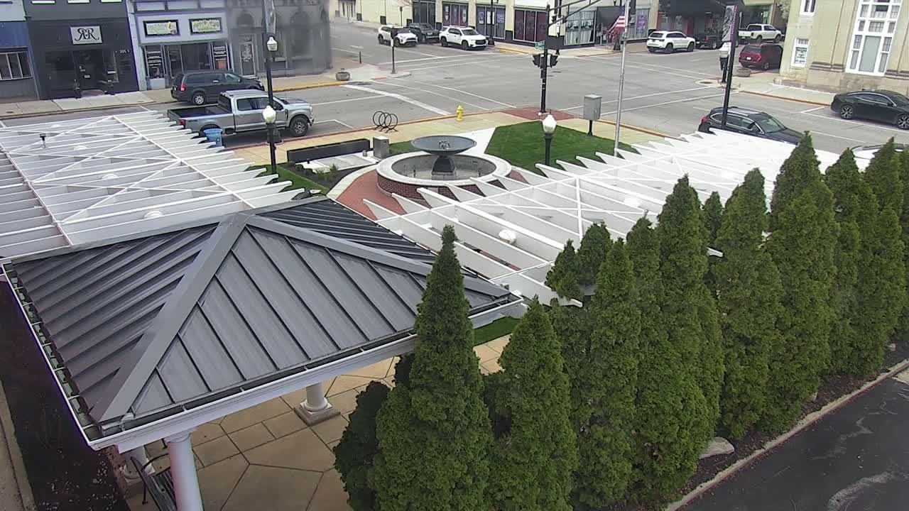 An aerial view shows a town square with a fountain, a white pergola structure, and a row of evergreen trees on a partly sunny day.