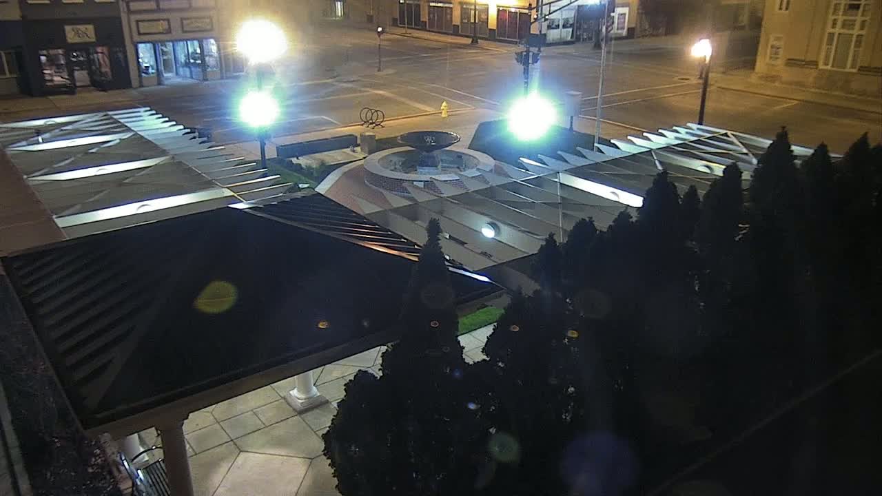 An overhead nighttime view of a town square featuring a fountain, illuminated by streetlights, surrounded by dark evergreen shrubs and a partially visible building with a dark roof.