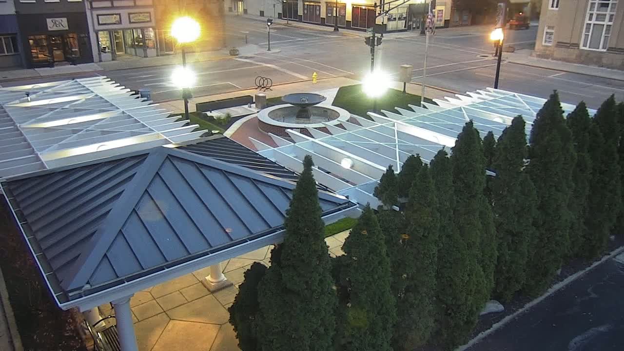 An overhead view shows a paved plaza with a fountain at its center, surrounded by evergreen trees and a modern metal-roofed structure with a glass canopy overhead, all under the soft light of dusk.