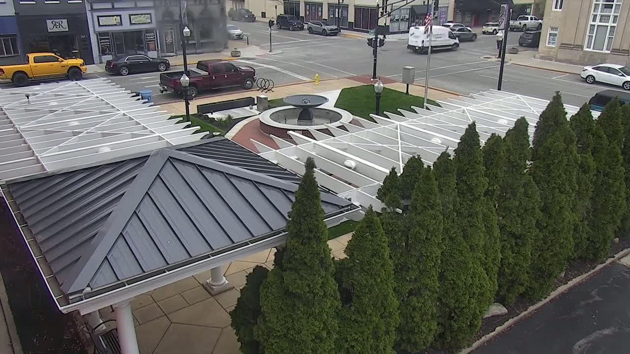 A dark gray roofed structure with a white pergola sits adjacent to a small fountain in a plaza surrounded by evergreen trees, with a street and parked cars visible in the background on a partly cloudy day.