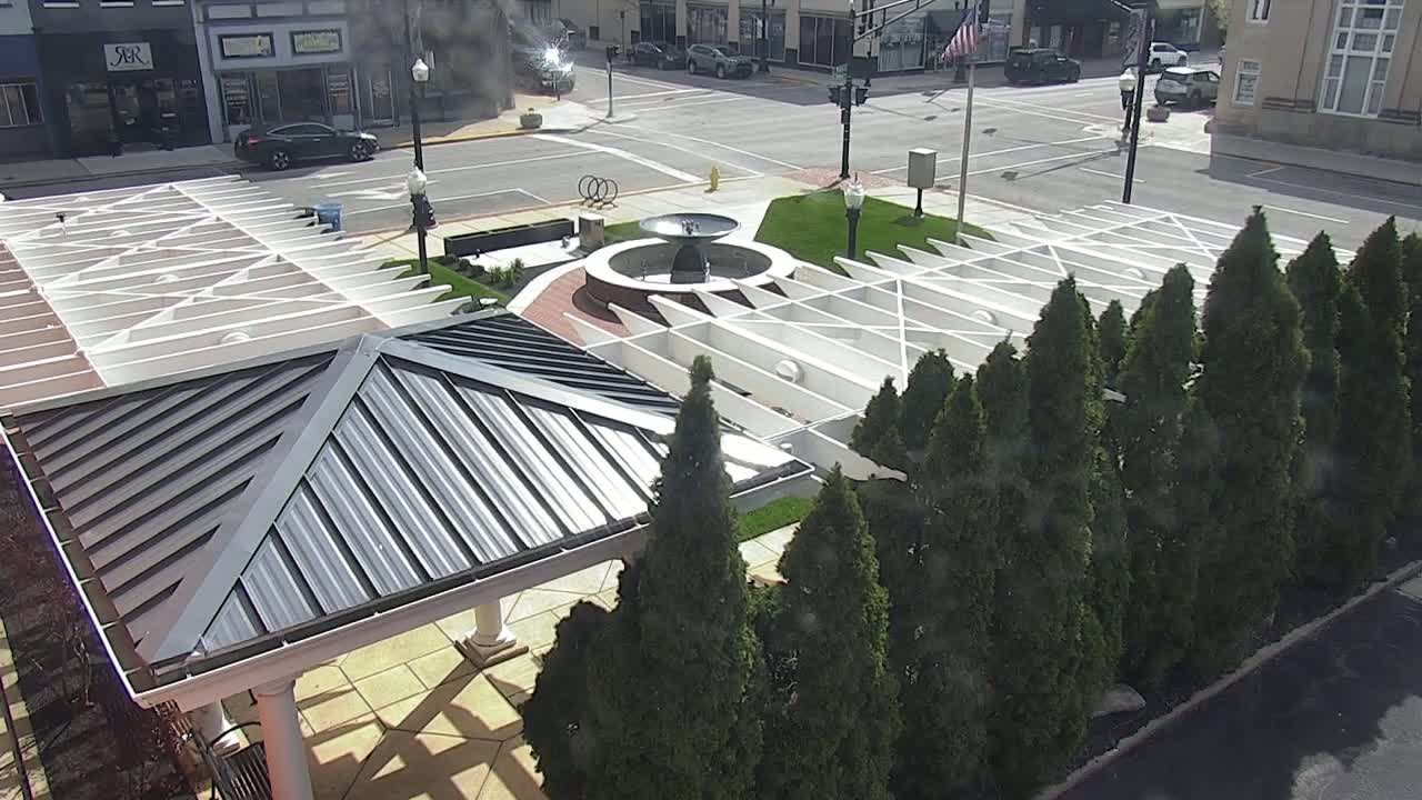 An aerial view of a town square featuring a fountain, a gazebo with a metal roof, and a row of evergreen trees on a sunny day.