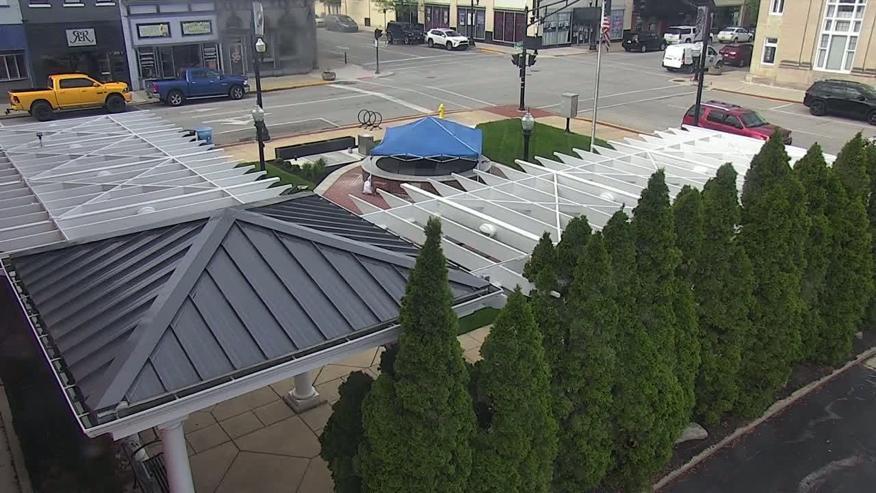 An overhead view shows a partially covered plaza with a blue tarp over a circular structure, surrounded by evergreen trees and a gray metal-roofed structure.