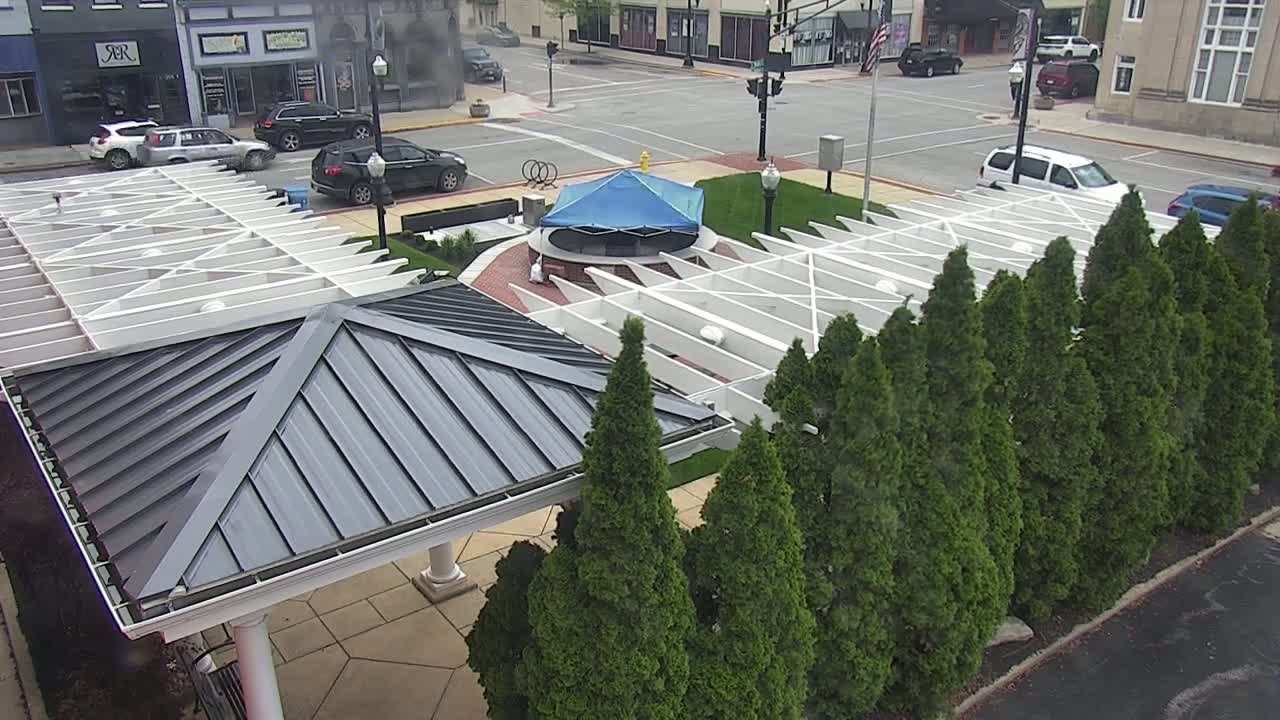 A partly cloudy day reveals a town square with a covered pavilion, a blue-covered structure in the center, and rows of evergreen trees lining the edges of a paved area.
