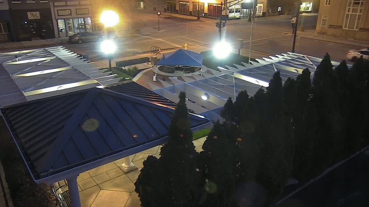 An elevated nighttime view shows a town square with a blue canopy, surrounding landscaping, and a modern pavilion with a dark blue roof.