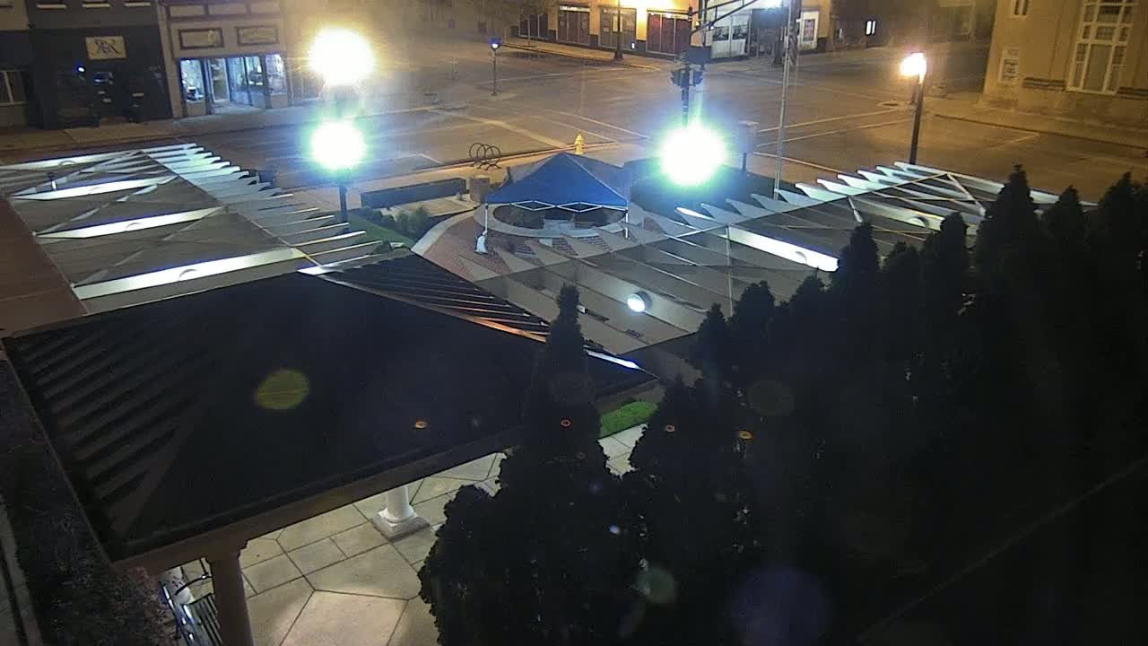An overhead nighttime view shows a plaza with a blue canopy and surrounding trees under bright streetlights.