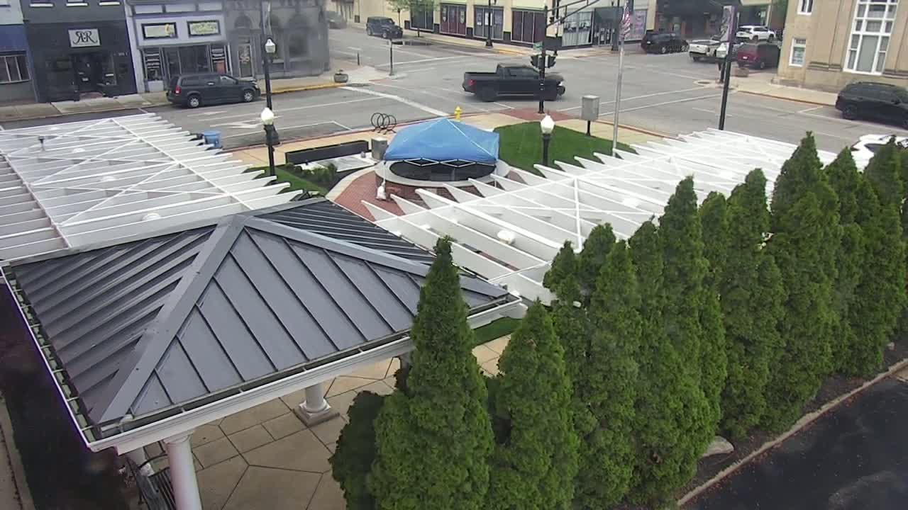 A paved area with white and gray pergolas, a covered structure, and a row of evergreen trees is situated in front of buildings under a partly cloudy sky.