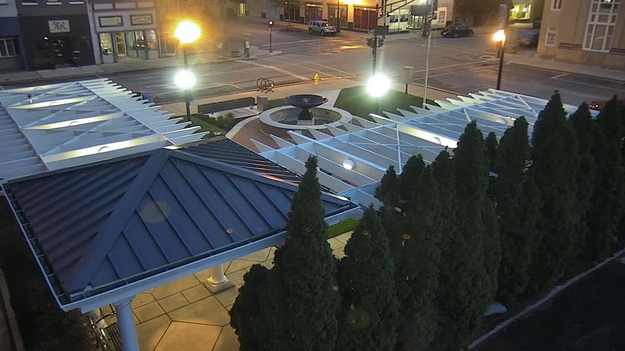 A nighttime aerial view shows a paved plaza with a central fountain, surrounded by evergreen trees and a pergola with a dark-blue roof.