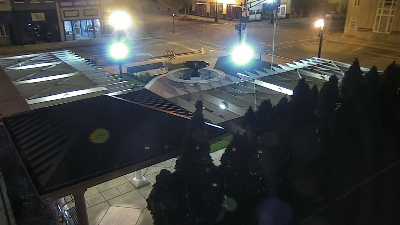 An illuminated nighttime view of a circular fountain surrounded by dark evergreen trees and a modern, angular pavilion.