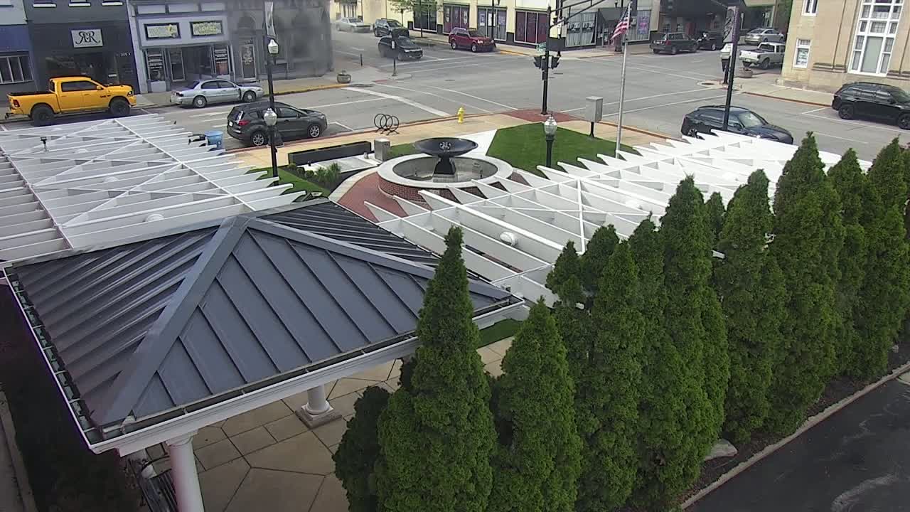 A dark-roofed pavilion and white pergola surround a central fountain in a town square, bordered by a row of evergreen trees on a partly cloudy day.
