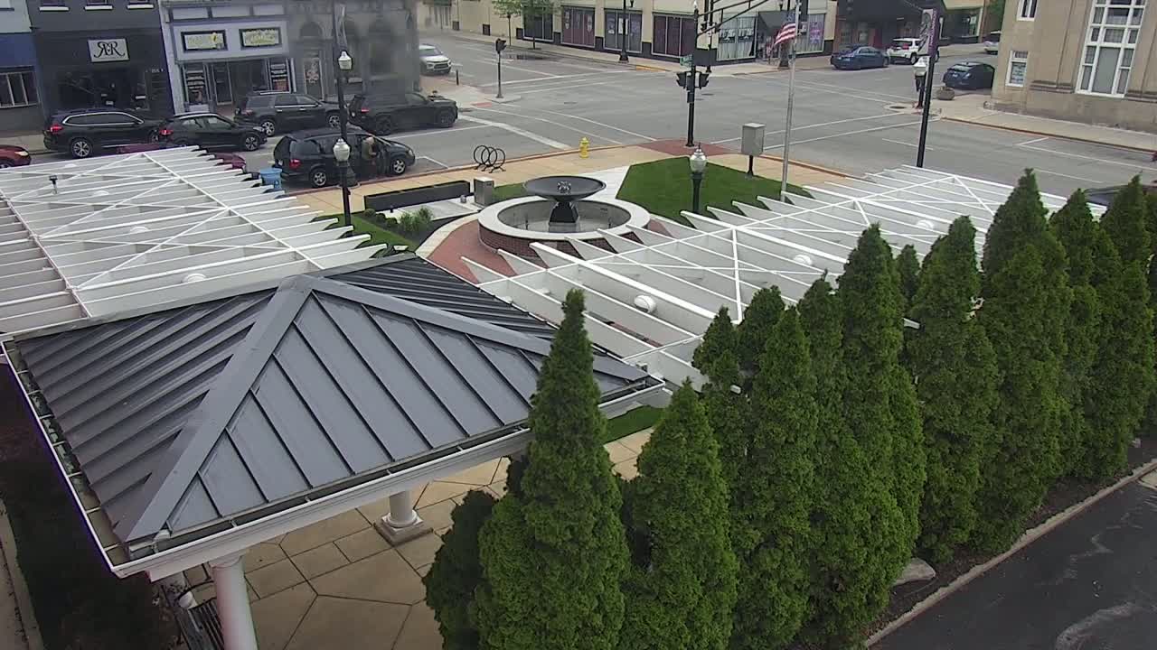 An overhead view shows a plaza with a central fountain, surrounded by evergreen trees and covered seating areas under white and gray metal structures, on a partly sunny day.