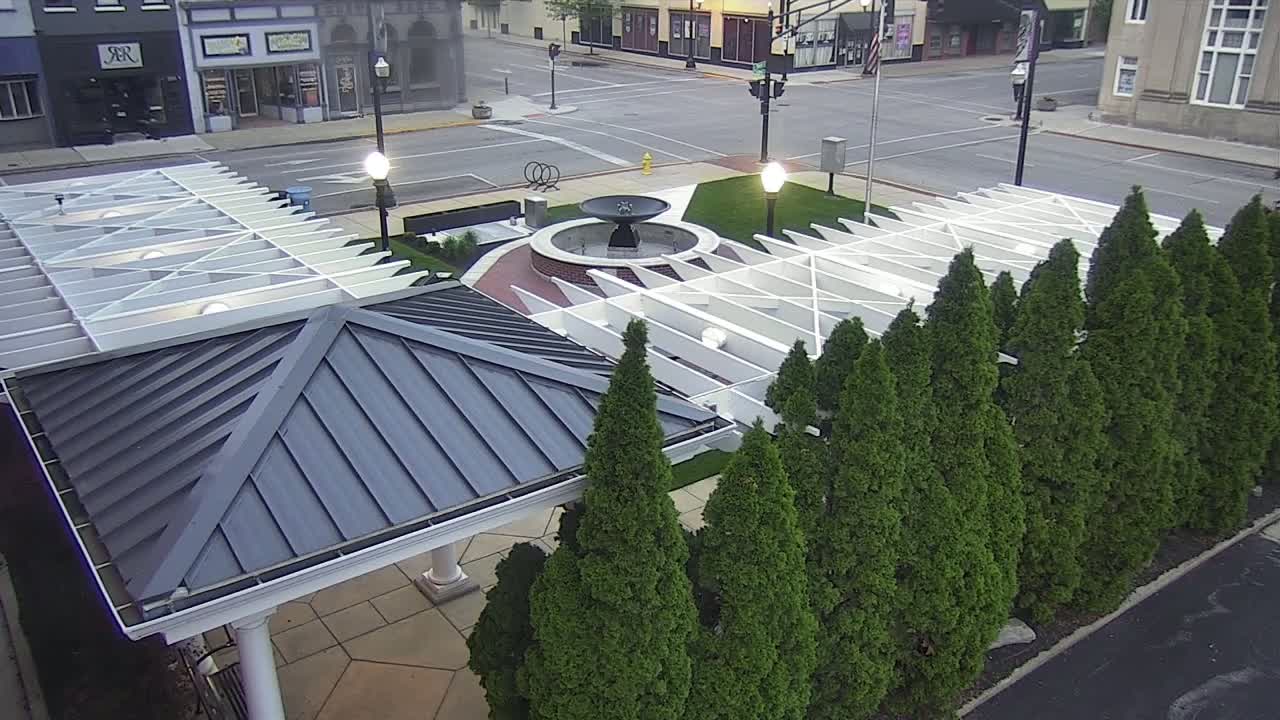 An aerial view shows a gazebo with a dark gray metal roof next to a white pergola structure surrounding a central fountain, all bordered by a row of evergreen trees on a partly sunny day.
